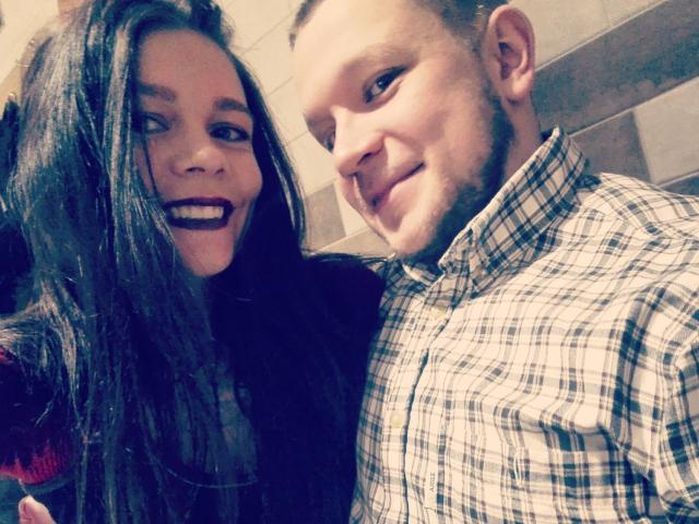 VulcanoX, con lunghi capelli scuri e rossetto scuro, sorride, ed è in piedi accanto a VulcanoX con i capelli corti e la barba, che indossa una camicia a quadri, entrambi sorridono..
