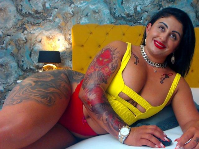 Luisahotass is a curvy Latina with tattoos, red lipstick, yellow cutout top, red panties, and a gold chain, lying on a bed with a yellow headboard.