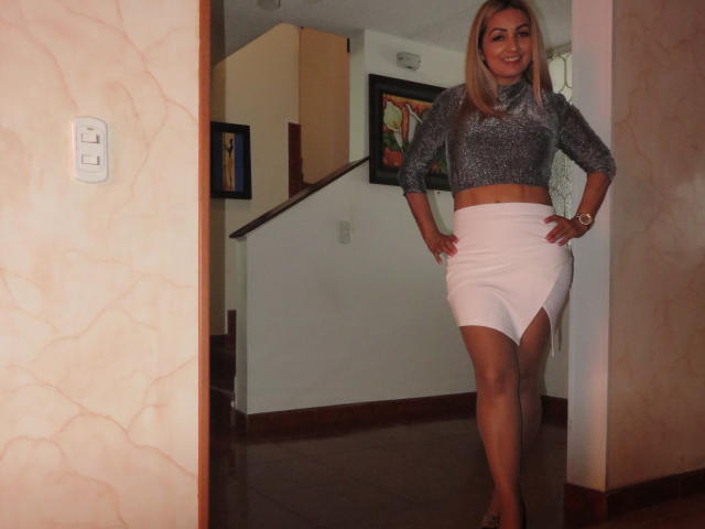 SexyBush, standing in a doorway, wearing a sparkly gray top and a tight white skirt with a high slit, showing her toned legs and smiling with hands on hips.