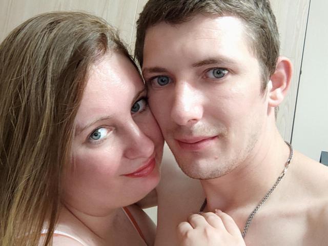JenAndPeter têm olhos azuis, os seios de JenAndPeter são pequenos, Peter está sem camisa, usando uma corrente, JenAndPeter tem cabelo castanho, Peter tem cabelo castanho curto, ambos estão sorrindo, JenAndPeter usa batom rosa..