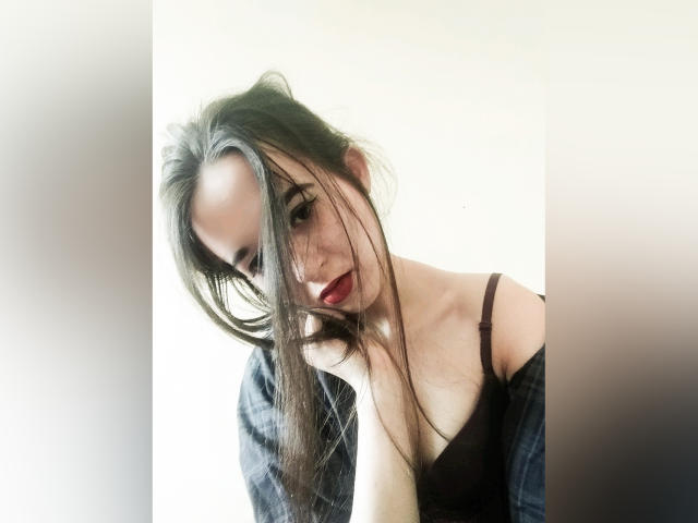 LuxCrow met lang bruin haar, rode lippenstift, een zwarte bh en een geruit overhemd met een open schouder, kijkt naar beneden, haar is lichtelijk rommelig over het gezicht, zacht licht, lichte schaduw aan de rechterkant..