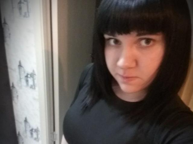 MaryCrystal met kort zwart kapsel, kijkt naar de camera, draagt een zwarte blouse, heeft kleine borsten, bleke huid, een serieuze uitdrukking, in een zwak verlichte kamer, foto..
