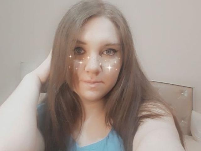 MaryCrystal a de longs cheveux bruns, des yeux bleus et une expression neutre. Elle porte un haut bleu avec des paillettes sur ses joues. Sa main est dans ses cheveux..