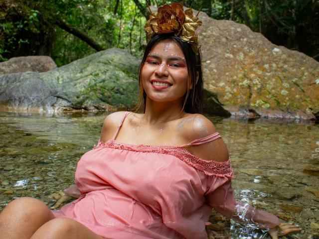 KorinYAmbar with wet skin, smiling, wearing a pink off-shoulder dress, sitting in a shallow stream, wearing a brown leaf headpiece, showing her big boobs and cute ass.