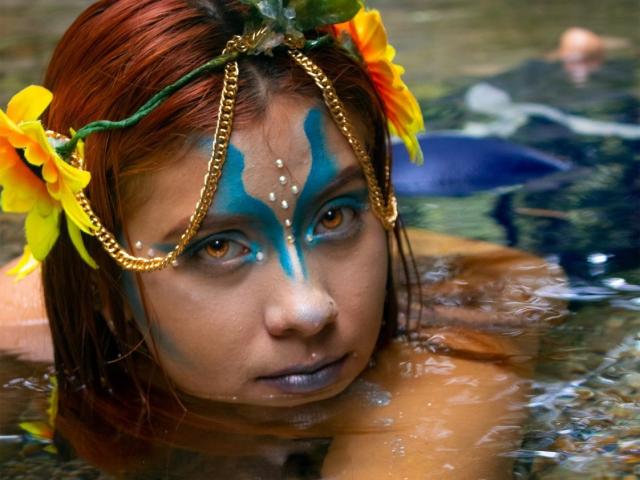 KorinYAmbar com cabelo vermelho molhado, maquiagem azul no rosto, adorno de cabeça com corrente dourada, flores amarelas e olhos castanhos na água, olhando para cima com expressão séria, mostrando um pequeno piercing no nariz..