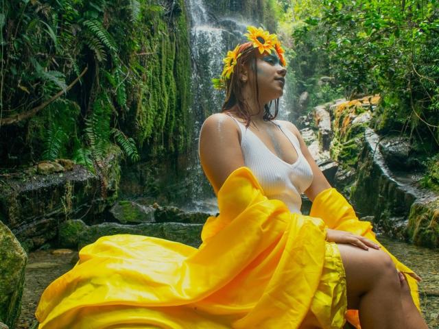 KorinYAmbar com uma coroa de flores amarelas, uma camiseta branca e um vestido amarelo, sentada perto de uma cachoeira, com a camiseta branca molhada, o vestido amarelo esvoaçante, água na pele, fundo de floresta, cabelo castanho molhado..