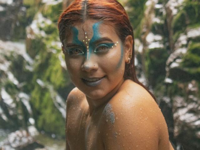 KorinYAmbar tem cabelos ruivos e molhados, maquiagem azul no rosto e batom cinza, com gotas de água em seus ombros nus, olhando para a câmera com um leve sorriso..