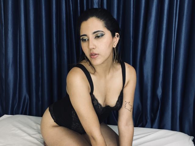 AgathaRose, avec ses cheveux foncés, sa lingerie en dentelle noire et ses boucles d'oreilles dorées, est agenouillée sur un lit, montrant ses seins et un petit tatouage sur son bras droit, et regarde la caméra de manière séduisante..
