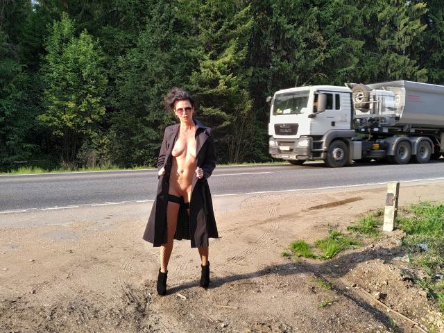 GannaWind stands on a dirt roadside, wearing an open black coat, no underwear, showing her tits and pussy, with black boots, and sunglasses, near a truck on a highway.