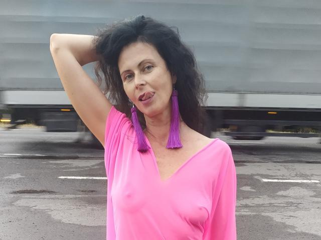 GannaWind with dark curly hair, pink off-shoulder top, visible nipples, purple tassel earrings, sticking tongue out, arm raised, blurred truck in background, standing on wet asphalt.