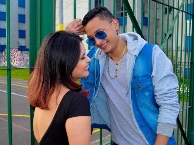 MelanyEtThomi com cabelo ombré em tons de vermelho-marrom, blusa preta, e MelanyEtThomi em colete azul, camisa cinza e óculos de sol estilo aviador azuis, sorrindo uma para a outra perto de uma cerca verde..