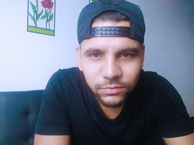 ShadowDiego with a black baseball cap, short beard, and black t-shirt, looking directly at the camera with a neutral expression. His brown eyes and medium skin are visible.