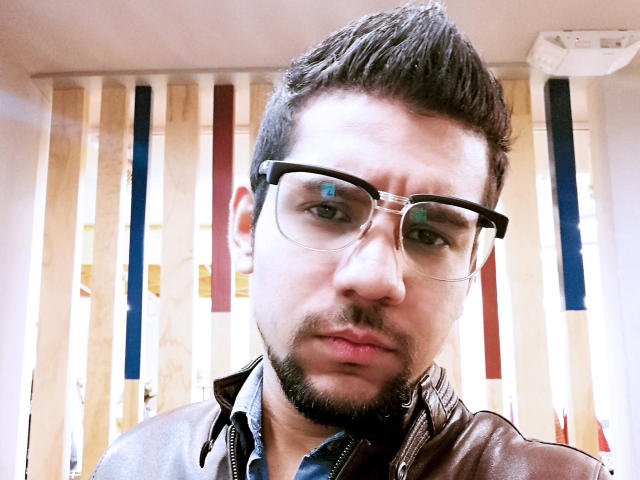 ShadowDiego has a goatee, black hair, and wears glasses with blue paint on them. He's in a brown leather jacket with a blue shirt underneath.