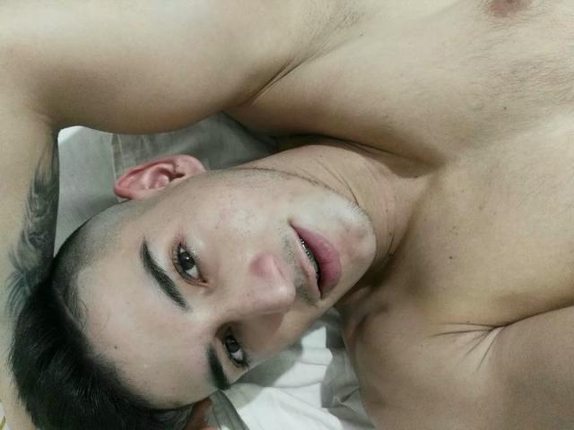 JonathanJeremiah, shirtless, lying on white bed, dark short hair, arched eyebrows, slight stubble, brown eyes, arms above head, subtle tattoo on left arm, soft light on face.