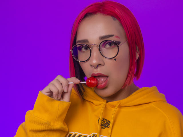 Lara Carter, con un corte de pelo rojo, gafas, piercing en la nariz y una sudadera amarilla, lamiendo una piruleta roja, fondo púrpura, lengua afuera, linda, intelectual, sexy, pequeña marca de nacimiento negra debajo del ojo derecho..