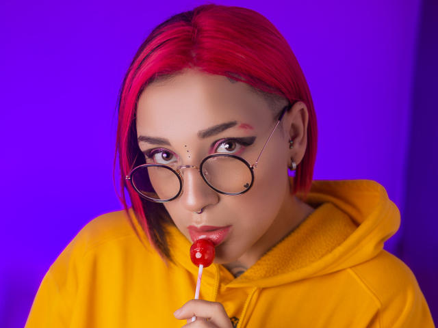 Lara Carter, avec des cheveux teints en rose vif, des lunettes rondes, un piercing au nez, un sweat à capuche jaune et un bonbon à la cerise, regardant la caméra, fond violet. Les seins ne sont pas visibles, le visage est en focus..