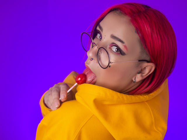 Lara Carter, met rood haar, een bril en een neusring, likt aan een rode lolly, draagt een gele hoodie, en de achtergrond is paars. Haar borsten zijn zichtbaar onder de hoodie..