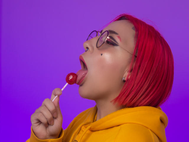 Lara Carter mit rotem Bob, gelber Kapuzenjacke und Brille, die eine rote Lutscher leckt, mit geschlossenen Augen und herausgesteckter Zunge, Nasenpiercing, lila Hintergrund..