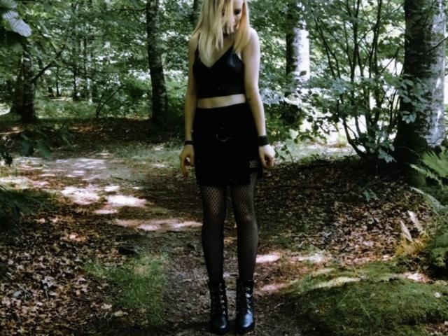 DreamArinna in piedi in una foresta, indossa un top corto in pelle nera, una minigonna nera, collant a rete e stivali neri con lacci, con i capelli biondi e un braccialetto al polso..