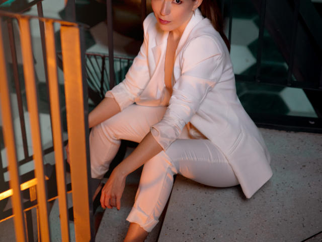 GingerLilli, sitting on a stair, wearing a white puffy jacket and white pants, with her legs crossed, looking at the camera, light brown hair, subtle smile.