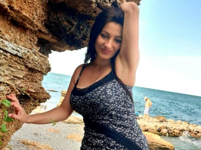 GinaONeon with dark hair, wearing a black and white floral dress, standing by rocky coastline, arm raised, smiling, showing her tits, with ocean and rocks in background.