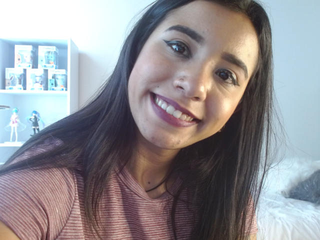 AshleyLover has long black hair, brown eyes, and a smiling face with pink lipstick. She's wearing a pink striped shirt, showing her cute nose and small tits.