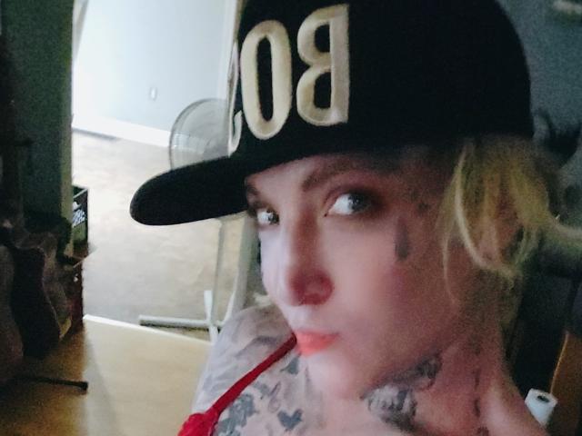 Madelinej, avec ses cheveux blonds, sa casquette de baseball noire et ses tatouages, regardant l'appareil photo avec des lèvres pincées, le bandeau de son soutien-gorge rouge visible, une peau pâle et des yeux légèrement flous..