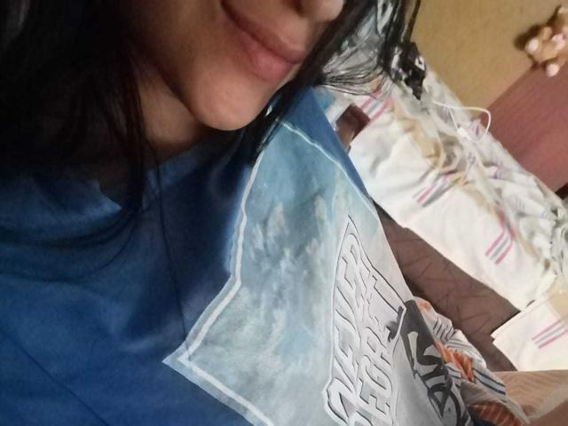 Samantha Sweett, mit einem leichten Lächeln, ihr dunkles Haar bedeckt teilweise ihr Gesicht, trägt ein blaues und weißes T-Shirt mit Aufdruck, ihre kleinen Brüste sind sichtbar, sie liegt auf einem Bett..