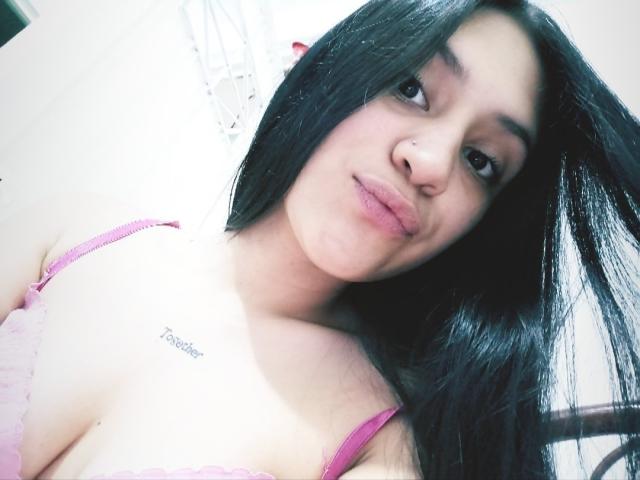 SamanthaSweett, com cabelos longos e pretos, sutiã de renda rosa, lábios carnudos, piercing no nariz, tatuagem no lado direito do peito com a palavra "Pooper". Sorriso suave, pele clara, seios de tamanho médio, selfie casual..