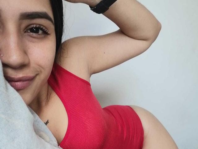 SamanthaSweett, con un pequeño piercing en la nariz, vestida con un traje de baño rojo de una sola pieza que muestra sus senos, y una pulsera negra, acostada sobre una manta gris, sonriendo ligeramente..