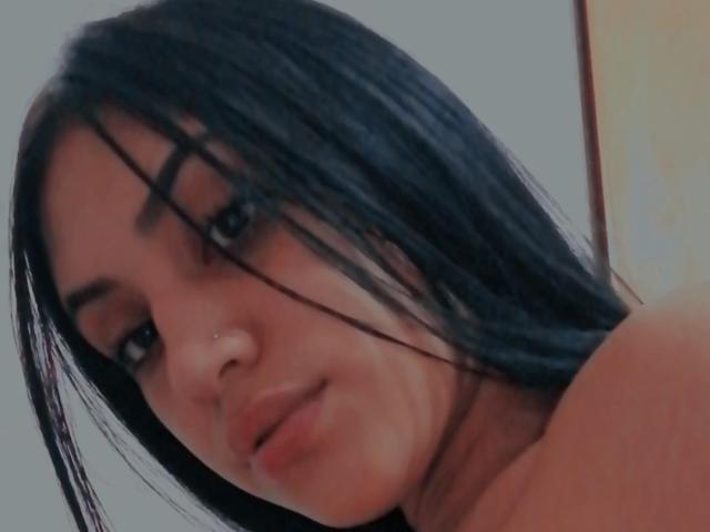 SamanthaSweett tiene el cabello negro largo que le cubre un ojo, un pequeño piercing en la nariz y una ligera sonrisa, con los hombros descubiertos. Está mirando a la cámara con una expresión relajada..