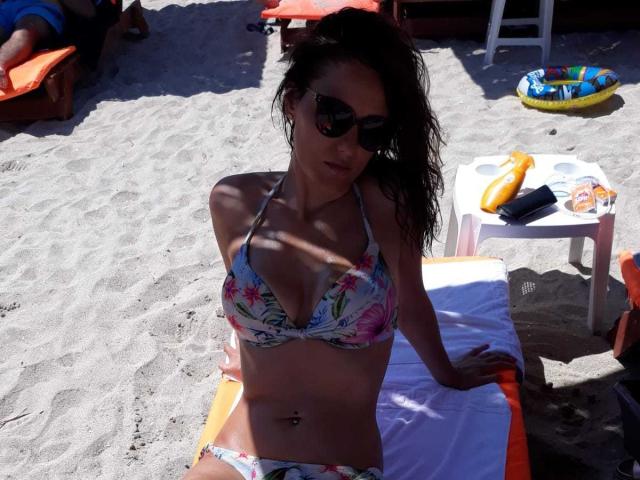 JoliDanielle in a floral bikini, sunbathing on a towel, with sunglasses on, and a belly button piercing, sitting on a sandy beach with a small white table nearby.