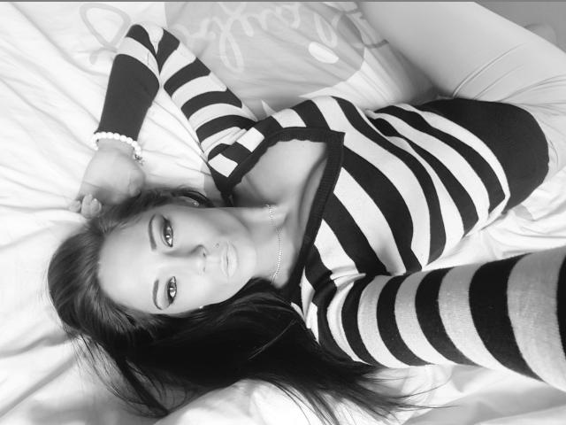 KaydenKross sdraiata su un letto, indossa un abito a righe nero e bianco, lunghi capelli scuri, occhi verdi, braccialetto al polso destro, guarda la fotocamera, luce soffusa, fotografia in bianco e nero..