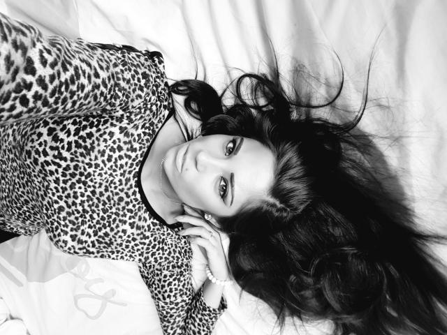 KaydenKross with long dark hair, wearing a leopard print top, lying on a bed, looking up with serious eyes, hands near her face, black and white photo.