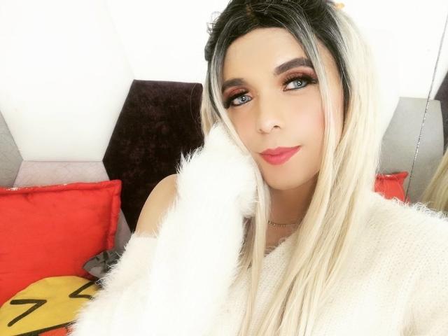 Kimberllys with long blonde and black hair, wearing a white fuzzy sweater, has red lipstick, and smoky eye makeup, smiling at the camera, showing a cute, sexy face.