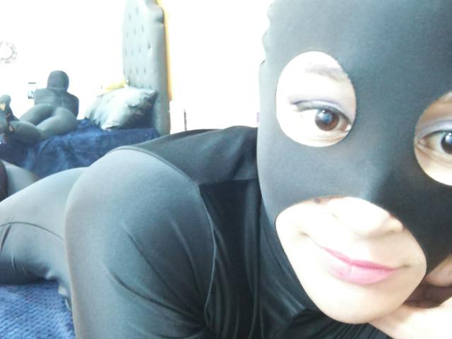 AllisonGag, portant un masque noir et une combinaison moulante, est allongée sur un lit bleu, souriant à la caméra avec ses yeux marrons, ses lèvres roses et son visage mignon. Ses fesses sont visibles..