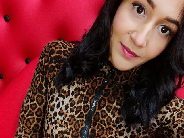 AlisonGag with dark curly hair, brown eyes, nose piercing, pink lipstick, wearing a leopard print zip-up top, sitting on a red tufted chair. Boobs partially visible.