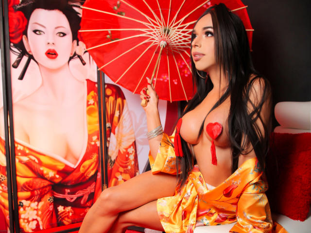 Jhovanna, met lang zwart haar, draagt een oranje zijden kimono, waarbij haar borsten zichtbaar zijn met rode hartvormige bedekking. Ze houdt een rode paraplu vast en zit naast een portret van zichzelf..