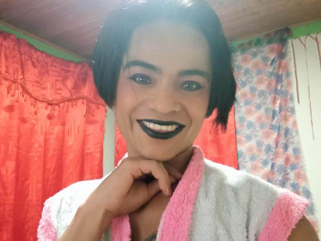 ChicoFemboy com cabelo preto curto, batom escuro e toalha rosa e branca, sorrindo, com a mão no queixo, olhando para a câmera. Cortinas vermelhas e florais ao fundo..