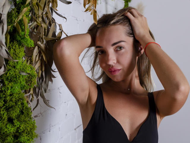 LettieSweet with hands in blonde hair, wearing a black v-neck, showing her tits, green moss, and brown leaves in the background, wearing a red bracelet.