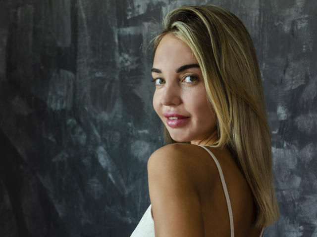 LettieSweet with blonde hair, green eyes, and pink lips, smiling at the camera, showing her white top and bare shoulders, looking sexy and cute, with a soft, natural look.