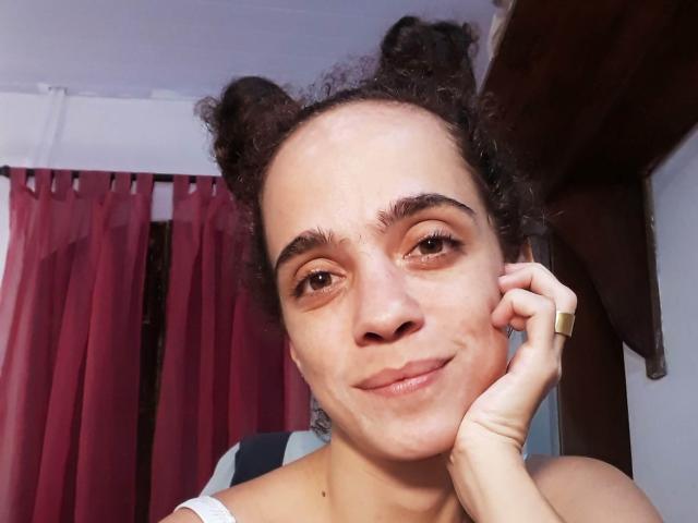 AmyFlores, avec des cheveux bouclés attachés en deux chignons, souriant légèrement, tenant un téléphone à l'oreille, portant un haut blanc, avec des rideaux rouges en arrière-plan, un anneau brun à l'annulaire, peau claire, yeux marrons..