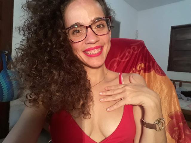 AmyFlores, con i capelli castani ricci, gli occhiali rossi e la canottiera rossa, sorride, mostrando il seno, con una mano sul petto, rossetto rosa, orologio da polso marrone e un asciugamano rosso sullo sfondo..