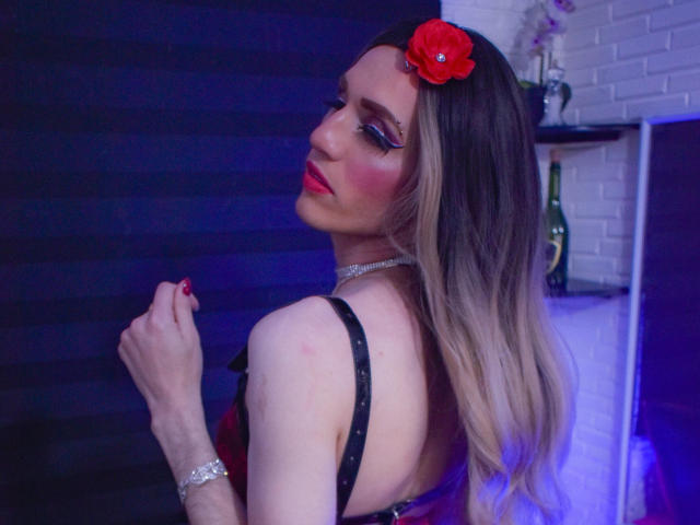 EvanghelineHof with long blonde hair, red flower in hair, black strap dress, red lipstick, blue eye makeup, bracelet, looking over shoulder, hand near chest, blue and white lighting.