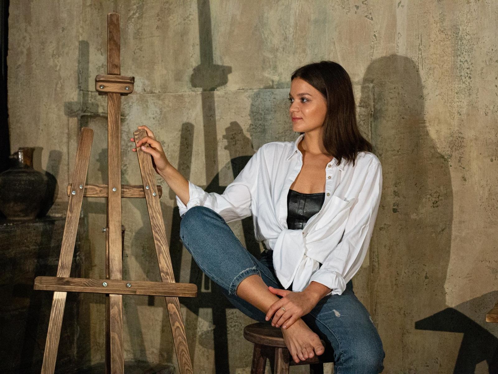 PennySmile, with brown hair, sits on a stool, legs crossed, wearing a white shirt, black top, and jeans, resting hand on wooden easel, looking to the side.