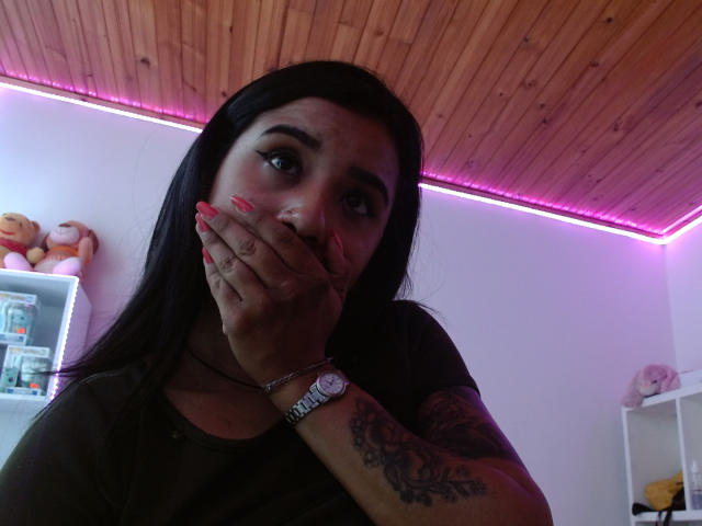 AshleyLover mit langem, schwarzem Haar, die sich mit ihrer Hand den Mund bedeckt, hat rosafarbene Nägel, ein Tattoo am rechten Arm und trägt ein dunkelgrünes Hemd..