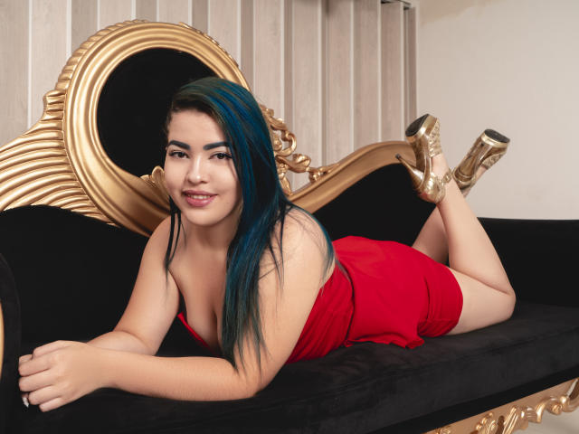 KimLafayette, met blauwe highlights, ligt op een zwarte fluwelen bank in een rode jurk, gouden laarzen, glimlachend, waarbij haar achterwerk zichtbaar is, en ze ziet er schattig uit..