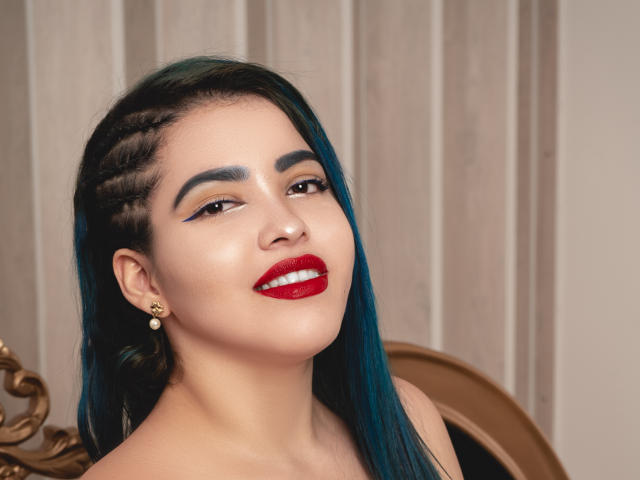 KimLafayette tem cabelo azul-esverdeado, sobrancelhas escuras, batom vermelho e um sorriso. Ela está usando um brinco dourado e tem o cabelo trançado em um dos lados. Seus seios não estão visíveis..
