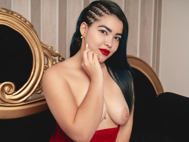 KimLafayette, con i capelli verdi intrecciati, rossetto rosso e orecchini, senza top, con piccoli seni, che si tocca il mento, indossa pizzo rosso, seduta su una sfarzosa sedia nera e dorata..
