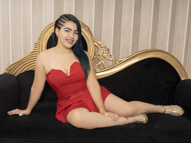 KimLafayette, una donna latina con riflessi blu, seduta su un divano di velluto nero con bordi dorati, indossa un abito rosso senza spalline, mostrando il seno e i glutei, sorride e indossa tacchi dorati..